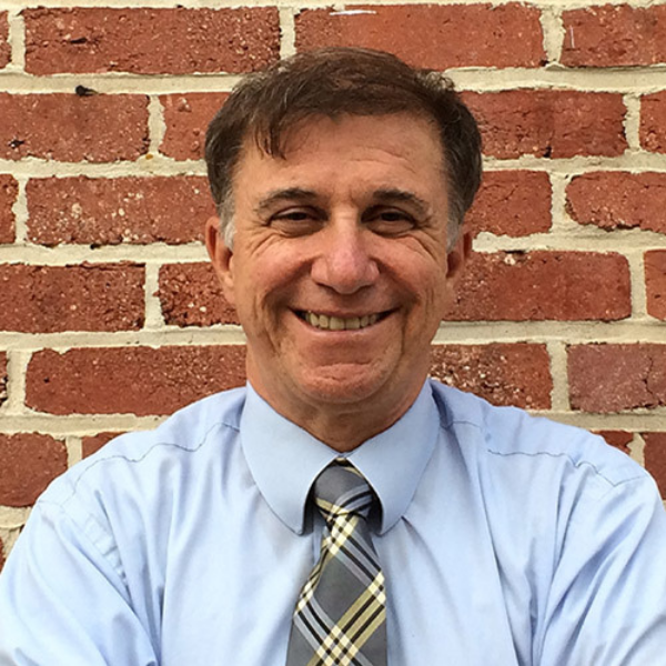 Vincent DeMarco in shirt and tie smiling at the camera in front of a brick wall